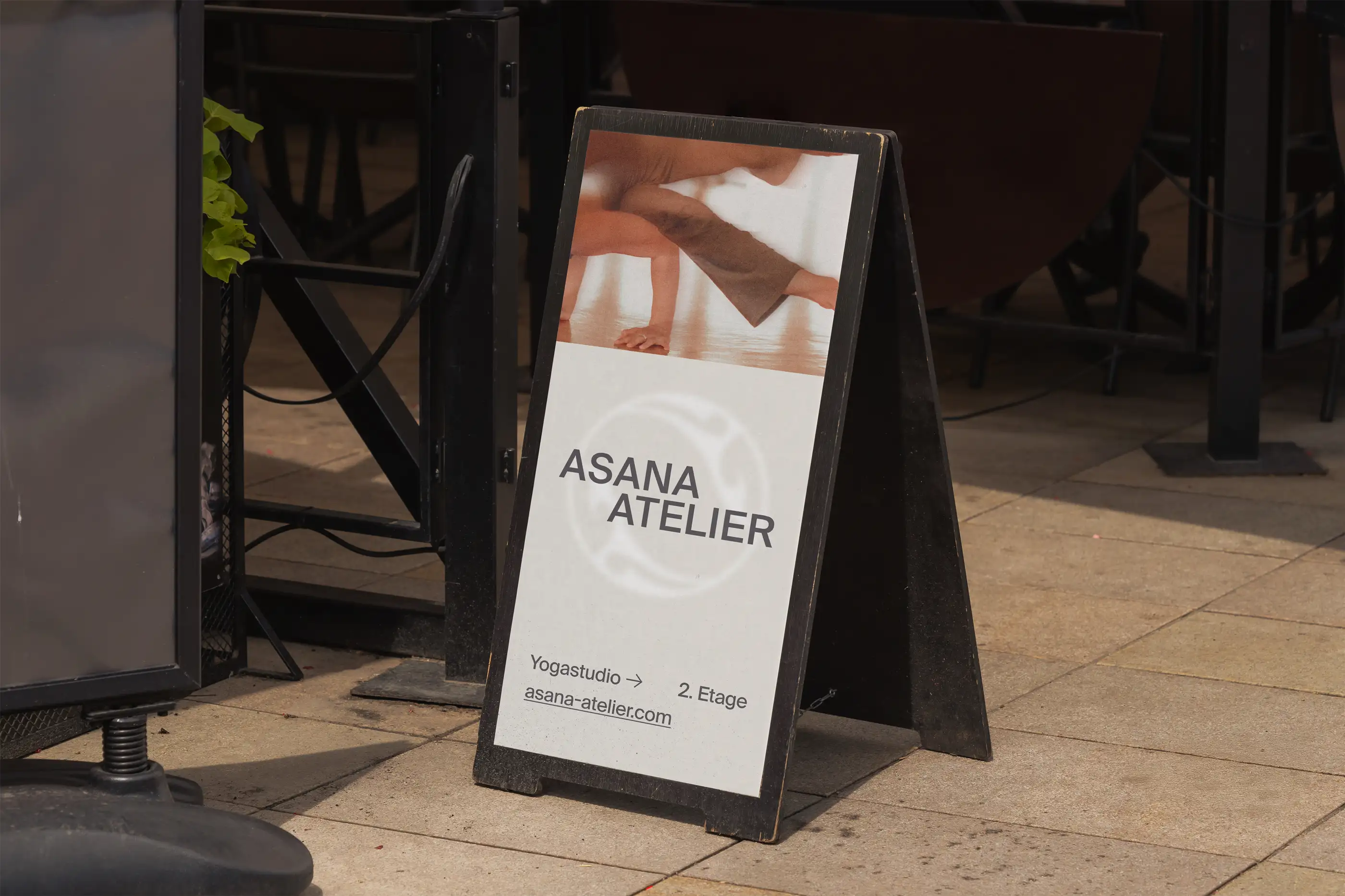 Schwarzes Werbeschild auf dem Bürgersteig wirbt für das Yogastudio Asana Atelier im 2. Stock und zeigt das Bild einer Person in einer Yogapose.